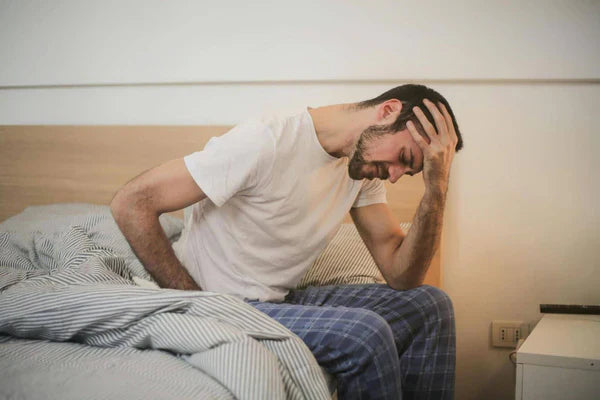 A man sitting on the edge of a bed, holding his head in discomfort 