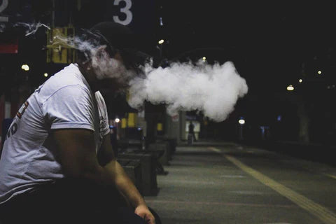 A black man sitting on a sidewalk bench vaping and puffing a dense cloud of nicotine smoke 