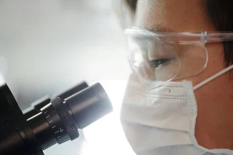 A medical worker with a mask and protective glasses looking down a microscope 