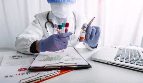 A medical examiner in protective hear sitting next to a laptop holding vials of drug testing samples 
